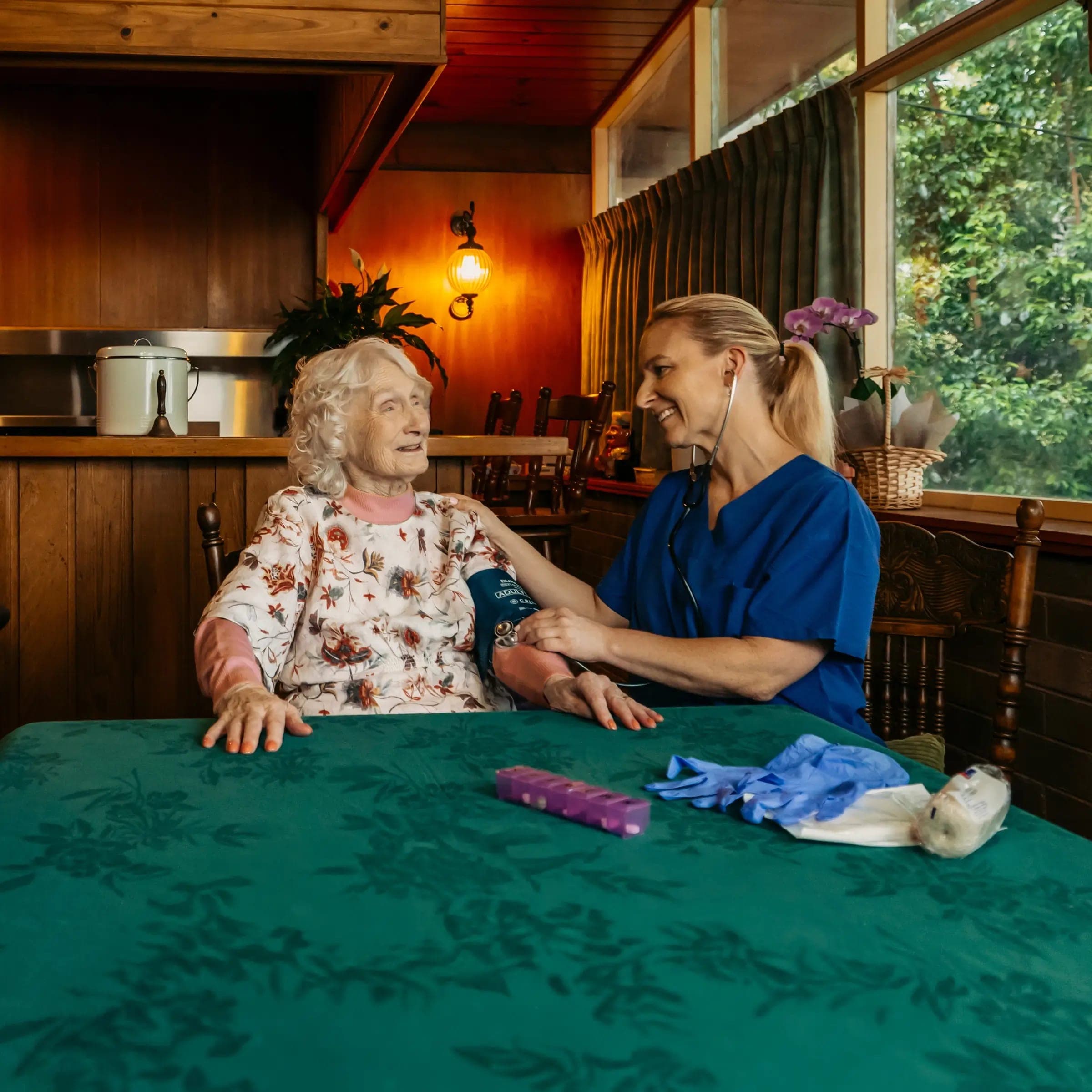 Registered Nurse takes a blood pressure reading for their older client at their dining table.